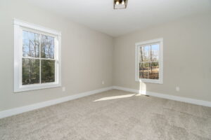 Empty room with beige walls and carpeted floor, featuring two windows with views of bare trees and a small light fixture on the ceiling.