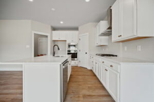 A modern kitchen with white cabinets, a central island featuring a built-in sink and dishwasher, stainless steel appliances, wood flooring, and light-colored walls.