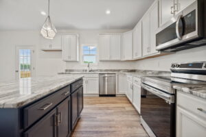 A modern kitchen with gray countertops, white cabinets, a large island with black cabinets, stainless steel appliances, a pendant light, and a window above the sink.