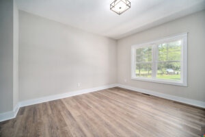A minimally furnished empty room with light gray walls, a large window with wood trim, and wood-look flooring. A modern ceiling light fixture hangs from the ceiling.