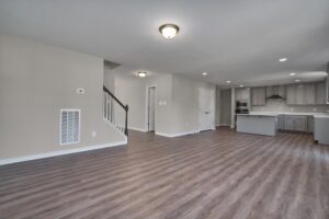 Spacious open-concept room with modern kitchen, laminate wood flooring, neutral-colored walls, and ceiling lights. Stairs with a black banister lead to the upper floor.