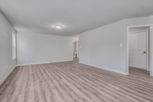 A spacious, empty room with light wood flooring, grey walls, a ceiling light, a window on the left, and a doorway leading to another room.