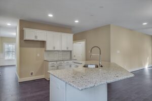 A modern kitchen with beige walls features white cabinets, a white tile backsplash, and a granite countertop island with a built-in sink and a sleek, curved faucet.
