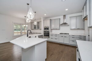 A modern kitchen features light gray cabinetry, a large island with a sink, stainless steel appliances, geometric pendant lights, and hardwood floors, with a view of an outdoor deck through the windows.