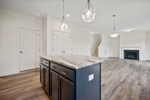 A modern, open-concept kitchen with a large island featuring a marble countertop, pendant lights, and dark cabinetry, leading into a living area with a fireplace and staircase.