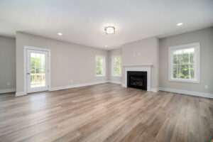 A spacious living room with wooden floors, light grey walls, large windows, and a fireplace. There is a door leading to an outdoor area on the left.