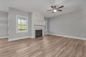 Spacious living room with light gray walls, a wooden floor, a ceiling fan, and a fireplace framed by two windows.