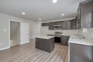 Modern kitchen with dark wood cabinets, stainless steel appliances, a central island, light granite countertops, and wood flooring. White doors are visible in the background.
