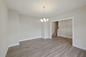 A minimalist room with light gray walls, wood flooring, a modern chandelier, and an open entryway leading to a staircase.