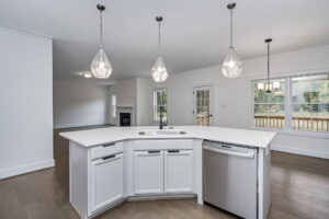 A modern kitchen with a white island featuring a sink and built-in dishwasher. Three geometric pendant lights hang above. In the background is the living area and a door leading outside.