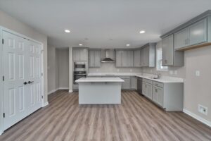 A modern kitchen with light gray cabinets, a central island, stainless steel appliances, wooden flooring, and recessed lighting.