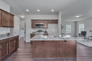 Modern kitchen with a central island featuring two bowls and cups on the countertop, stainless steel appliances, wooden cabinets, and an adjacent living area with a sitting chair and coffee table.