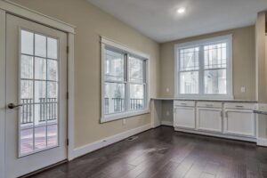 A bright room with dark hardwood flooring, white cabinets, large windows, and a glass door leading to a porch.
