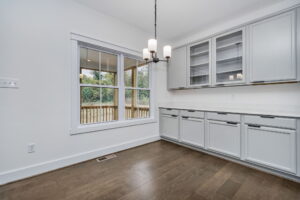 A kitchen area with wooden floors, gray cabinets, a chandelier light fixture, a large window overlooking a deck, and a white countertop beneath glass-fronted cupboards.