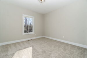Empty room with gray walls, a single window letting in natural light, and beige carpeted flooring.