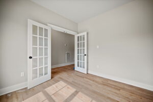 Empty room with light wood flooring, gray walls, and French doors. Sunlight casts shadows on the floor. A doorway leads to another space.
