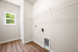 A clean, empty laundry room with beige walls, wooden flooring, and wire shelving above a small pet door, with a view of a window and a closed door.