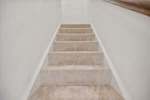 A beige carpeted staircase with white baseboards and a wooden handrail on the right side, leading upwards between clean white walls.