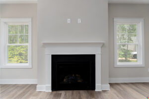 A modern fireplace with a white mantel is centered between two windows in a room with light gray walls and wood flooring.