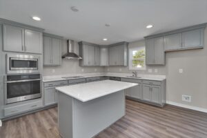 A modern kitchen featuring gray cabinets, stainless steel appliances, a large island with a white marbled countertop, and a window above the sink. The floor is dark wood.