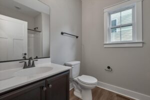 A bathroom with a sink, faucet, mirror, toilet, and a window. Walls are painted light gray. The floor is wooden, and there's a white door leading to a shower area.
