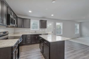 A modern kitchen with dark cabinetry, granite countertops, an island in the center, stainless steel appliances, and wooden flooring. Bright natural light enters through multiple windows.