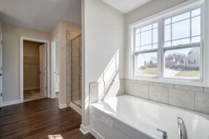 Modern bathroom interior with a large bathtub by a window, hardwood floors, and a glass shower door.