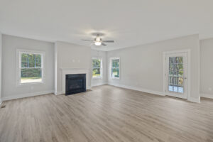 A spacious, empty living room with light wood flooring, white walls, a ceiling fan, a fireplace, three windows, and a glass door leading to an exterior balcony.