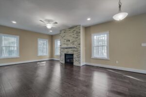 A spacious, unfurnished living room with dark hardwood floors, beige walls, white trim, a stone fireplace, three windows, a ceiling fan, and a hanging light fixture.