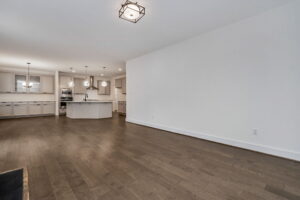 Modern open-concept kitchen and living area with hardwood floors, white walls, and recessed lighting. The kitchen features an island, stainless steel appliances, and pendant lights.
