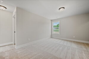 A carpeted, empty room with light beige walls, a window with a view of greenery, a closet door, and two ceiling light fixtures.