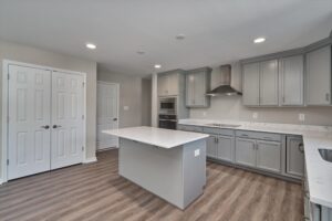 Modern kitchen with gray cabinets, stainless steel appliances, a center island with a light countertop, wood flooring, and recessed lighting. Two white double doors are visible in the background.