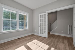 A room with large windows, French doors, light-colored walls, and hardwood flooring, adjacent to a hallway with a staircase. Plenty of natural light enters through the windows.