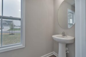 A small bathroom with a pedestal sink, round mirror, and a window showing a view of the outside. The walls are painted light gray.