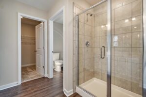 Modern bathroom with glass shower enclosure, beige tiles, and adjacent walk-in closet with open door.