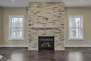 Living room with a stone accent wall featuring a fireplace in the center, flanked by two windows. The floor is dark wood, and the walls are painted beige.