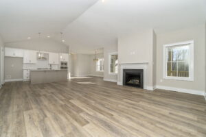 Modern, spacious living room with kitchen area featuring white cabinets, hardwood floors, a fireplace, and large windows.