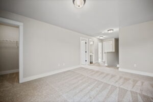 An empty carpeted room with beige walls, two ceiling lights, a walk-in closet on the left, and a bathroom door and countertop visible on the far side.