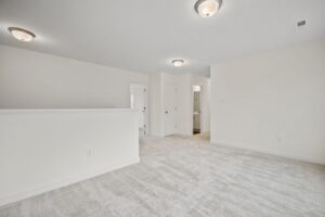 A spacious, empty room with white walls and ceiling lights. The floor is covered in light gray carpet. There are two doors, one of which leads to a small bathroom.