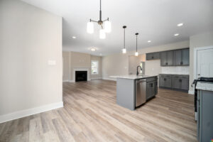 Modern kitchen and living area with light wood flooring, grey cabinets, central island with sink, pendant lighting, and a fireplace in the background.