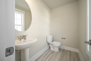 A modern, small bathroom with a white pedestal sink, toilet, and round mirror beside a window.