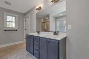 Modern bathroom with a large mirror, double sink vanity with blue cabinets, wall-mounted light fixtures, a walk-in shower with glass doors, and two windows providing natural light.