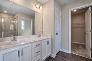 A modern bathroom featuring a double vanity with white cabinets and marble countertop, large mirror, glass shower enclosure, and doors leading to a closet and hallway.