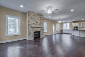 Spacious open-concept living area with dark hardwood floors, a stone fireplace, ceiling fan, and large windows. The space extends into a modern kitchen with white cabinetry and stainless steel appliances.