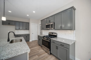 A modern kitchen featuring gray cabinets, granite countertops, a large island with a sink, stainless steel appliances, and a white subway tile backsplash.