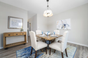 A dining room with a wooden table set for four, surrounded by upholstered chairs. The room features a sideboard, framed artwork on the walls, and a modern light fixture above the table.