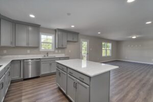 A modern kitchen with gray cabinets, stainless steel appliances, and a white marble island opens into a spacious living area with wood flooring and large windows.