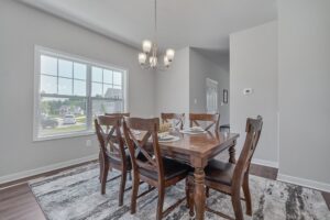A modern dining room featuring a wooden table set for four, large windows with street view, gray walls, and a pendant light.