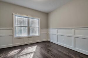 A small, empty room with beige walls, white wainscoting, a large double window, and dark hardwood floors.