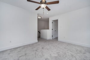 A room with light gray carpet, white walls, a ceiling fan with lights, and a white door partially open to a small hallway.